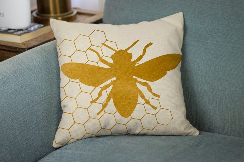 Puede incluir: Una almohada con una silueta de abeja dorada sobre un fondo blanco con un patr&oacute;n de panal.
