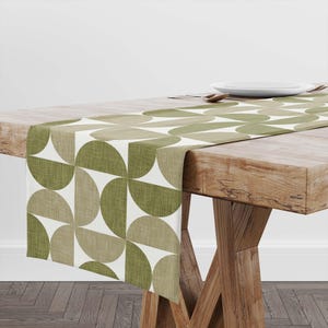 May include: A table runner with a geometric pattern of olive green and tan half-circles on a white background. The runner is displayed on a wooden table, showcasing its design and texture. The runner's dimensions are not visible.