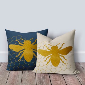 Puede incluir: Dos almohadas decorativas con un patr&oacute;n de panal y una silueta de abeja dorada. La almohada de la izquierda es azul marino y la almohada de la derecha es crema.