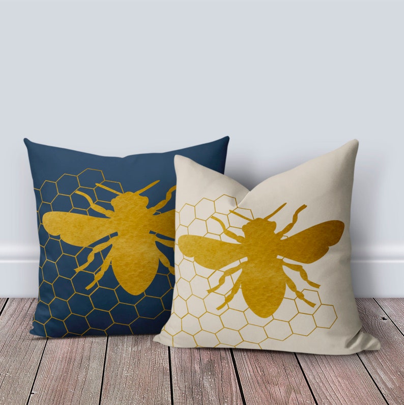 Puede incluir: Dos almohadas decorativas con un patr&oacute;n de panal y una silueta de abeja dorada. La almohada de la izquierda es azul marino y la almohada de la derecha es crema.
