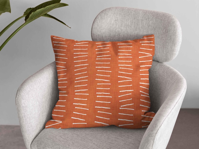 Puede incluir: Un coj&iacute;n cuadrado con una base de tela color naranja quemado y un patr&oacute;n repetitivo de columnas verticales compuestas por l&iacute;neas blancas horizontales cortas y discontinuas. El coj&iacute;n tiene una textura tejida visible y est&aacute; colocado.