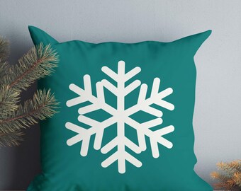May include: A square throw pillow in a solid teal color featuring a large, centered white snowflake graphic. The pillow has a smooth fabric texture and is positioned with green pine needles visible on the left and right sides of the frame.