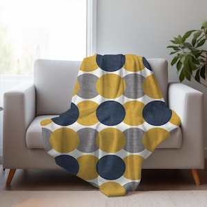 Puede incluir: Una manta con un patrón de círculos azul marino, amarillo mostaza y gris sobre un fondo blanco, colocada sobre un sofá blanco. El sofá tiene patas de madera y está situado cerca de una ventana. El diseño es repetitivo.