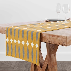 May include: A yellow and gray table runner with a geometric pattern. The runner is on a wooden table with two wine glasses and a plate.