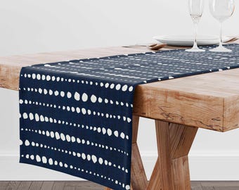 May include: A navy blue table runner featuring a repeating pattern of white dots. The runner is displayed on a wooden table, with place settings visible in the background. The design adds a touch of elegance to any dining setting.