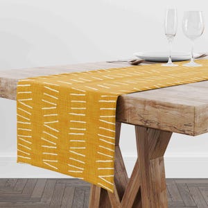 May include: Mustard yellow table runner with a white dash pattern. The runner is draped over a wooden table, with place settings and glassware visible in the background. The table has a natural wood finish.