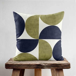 May include: A square throw pillow featuring a geometric pattern of navy blue and olive green semi-circles arranged on a white background. The fabric has a visible woven texture, creating a clean, repeating design across the entire surface.