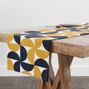 May include: A table runner with a geometric pattern of navy blue and mustard yellow semi-circles on a white background. The runner is displayed on a wooden table, showcasing its design and texture. The table runner is a decorative home decor item.