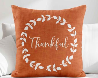 May include: A square throw pillow with a burnt orange fabric cover featuring a white printed wreath design. The word Thankful is written in a white cursive script in the center of the wreath. The fabric has a visible woven texture throughout.