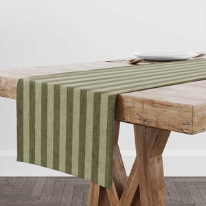 May include: A striped table runner in shades of olive green and beige, draped over a wooden table. The runner features vertical stripes and adds a touch of rustic elegance to the dining setting. A plate and cutlery are visible.