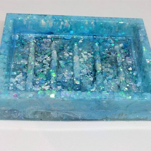 May include: A rectangular soap dish made of translucent blue resin. The dish is filled with iridescent glitter and has five raised bars. The edges of the dish are slightly rounded, and the overall effect is sparkly and decorative.