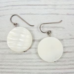 Peut inclure: Une paire de boucles d'oreilles rondes en nacre blanche avec des crochets argentés. Les boucles d'oreilles ont une surface texturée.