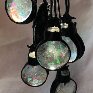 May include: A collection of round, iridescent pendants with black leather accents. Each pendant features a clear, glass-like orb filled with colorful, fractured pieces, suspended from a black cord. The orbs reflect light, creating a shimmering effect.
