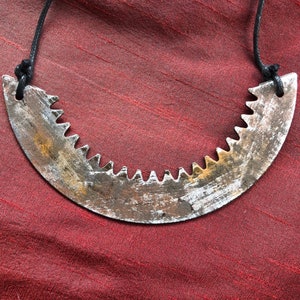 May include: A unique necklace featuring a curved, saw-blade pendant with jagged teeth. The metal has a distressed, weathered appearance with shades of silver, brown, and rust. The pendant is suspended from a black cord.