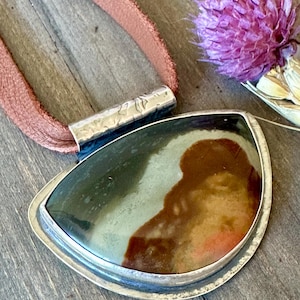 May include: A silver pendant necklace with a brown, green, and orange agate stone set in a silver bezel. The necklace is on a brown leather cord.