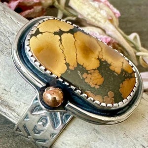 May include: A silver ring with an oval-shaped stone featuring a mix of brown and dark green patterns. The ring has a textured band and a small copper-colored accent. The stone is framed by a silver border.