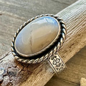 May include: Oval-shaped silver ring with a large, light-colored stone. The stone has a marbled appearance. The ring has a twisted rope-like border around the stone and a patterned band. The ring is displayed on a piece of wood.