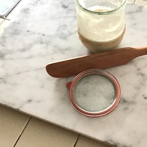 May include: A white marble cutting board with a glass jar of sourdough starter, a wooden spatula, and a glass lid with a red rubber seal.