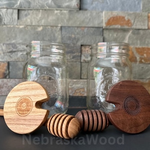 May include: Two glass jars with wooden lids and honey dippers. The lids are engraved with a circular design. One lid is light brown and the other is dark brown. The dippers are made of wood and have a honeycomb pattern.
