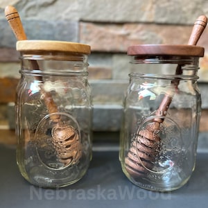 May include: Two glass jars with wooden lids and wooden honey dippers inside. The jars are clear glass with a textured surface. The lids are made of different types of wood, one is a light wood and the other is a darker wood. The dippers are both made of wood and have a rounded spoon shape.
