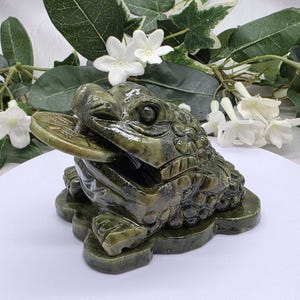May include: A dark green stone sculpture of a three-legged toad with a textured, bumpy back and a smooth underside. The toad has a wide mouth holding a flat, round coin with etched markings, and it is positioned on a flat, irregular base.