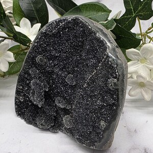 May include: A large, dark gray geode with a sparkling, crystalline surface. The geode is irregularly shaped and appears to be a natural stone formation. White flowers and green leaves are arranged behind the geode.