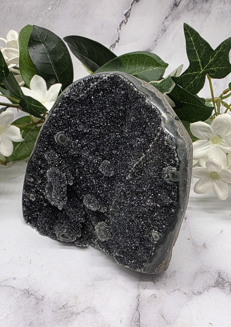 May include: A large, dark gray geode with a sparkling, crystalline surface. The geode is irregularly shaped and appears to be a natural stone formation. White flowers and green leaves are arranged behind the geode.