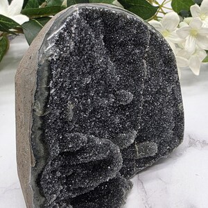 May include: A large, dark gray geode with a sparkling, crystalline interior. The geode has a rough, textured exterior and is displayed against a backdrop of white flowers and green foliage. A natural home decor item.