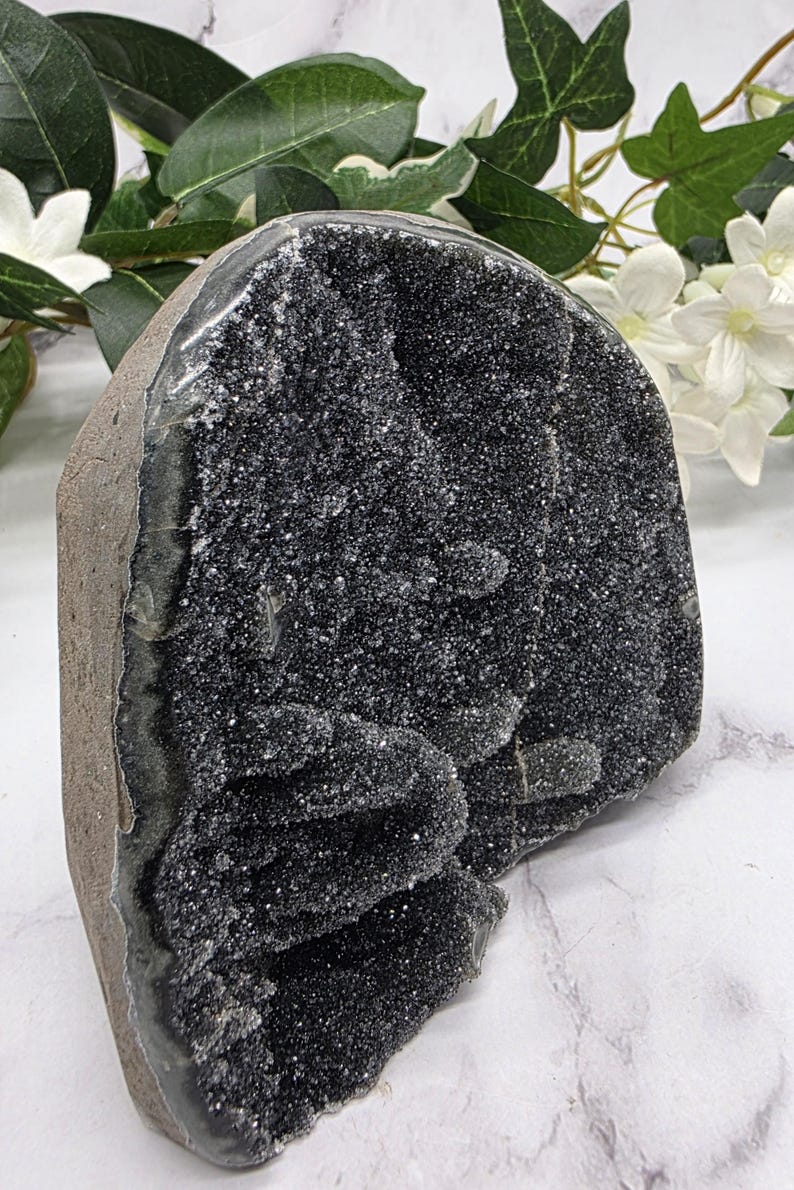 May include: A large, dark gray geode with a sparkling, crystalline interior. The geode has a rough, textured exterior and is displayed against a backdrop of white flowers and green foliage. A natural home decor item.