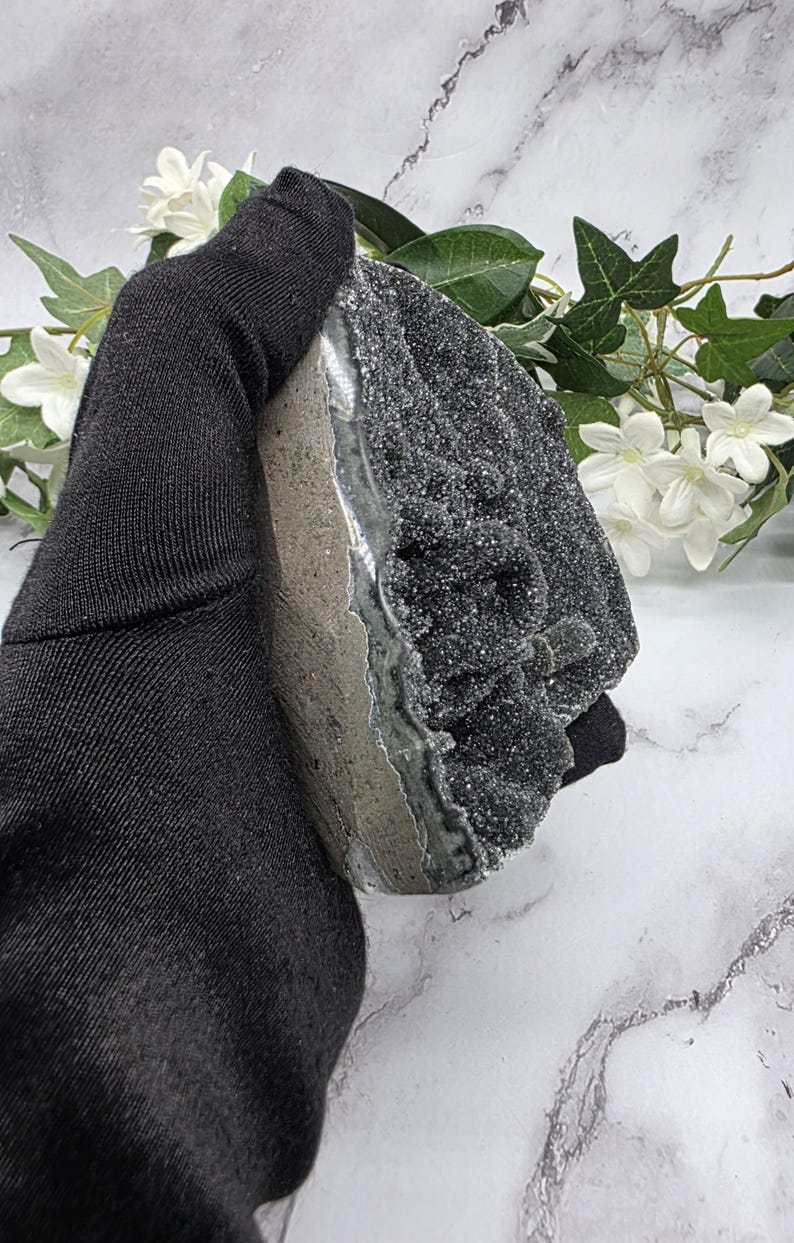 May include: A dark gray geode with a sparkling, crystalline surface. The geode is held by a black glove, showcasing its rough, textured exterior and natural color variations. White flowers and greenery are in the background.
