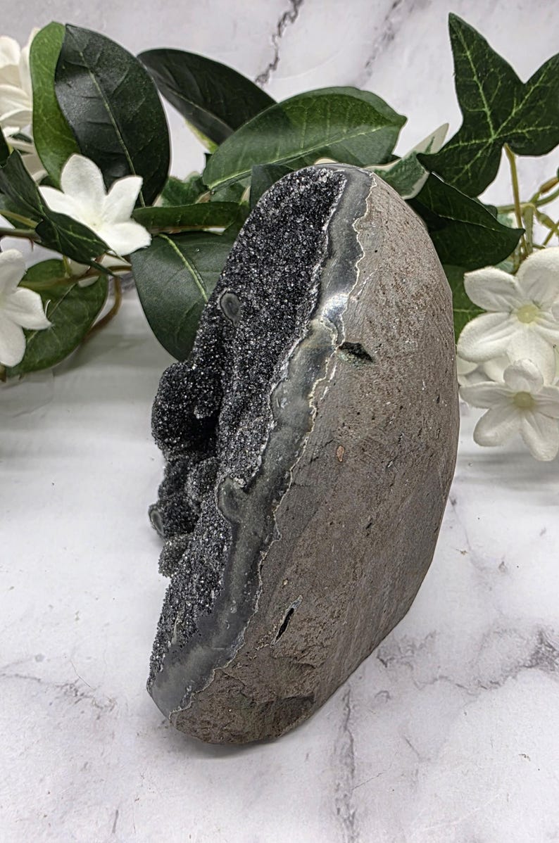 May include: A dark gray geode with a sparkling, crystal-like interior. The geode has a rough, textured exterior and a smooth, polished edge. White flowers and green leaves are in the background.