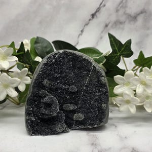 May include: A dark gray crystal geode with a sparkling, textured surface. The geode has a rounded shape with a rough, uneven texture. White flowers and green leaves are arranged around the geode.