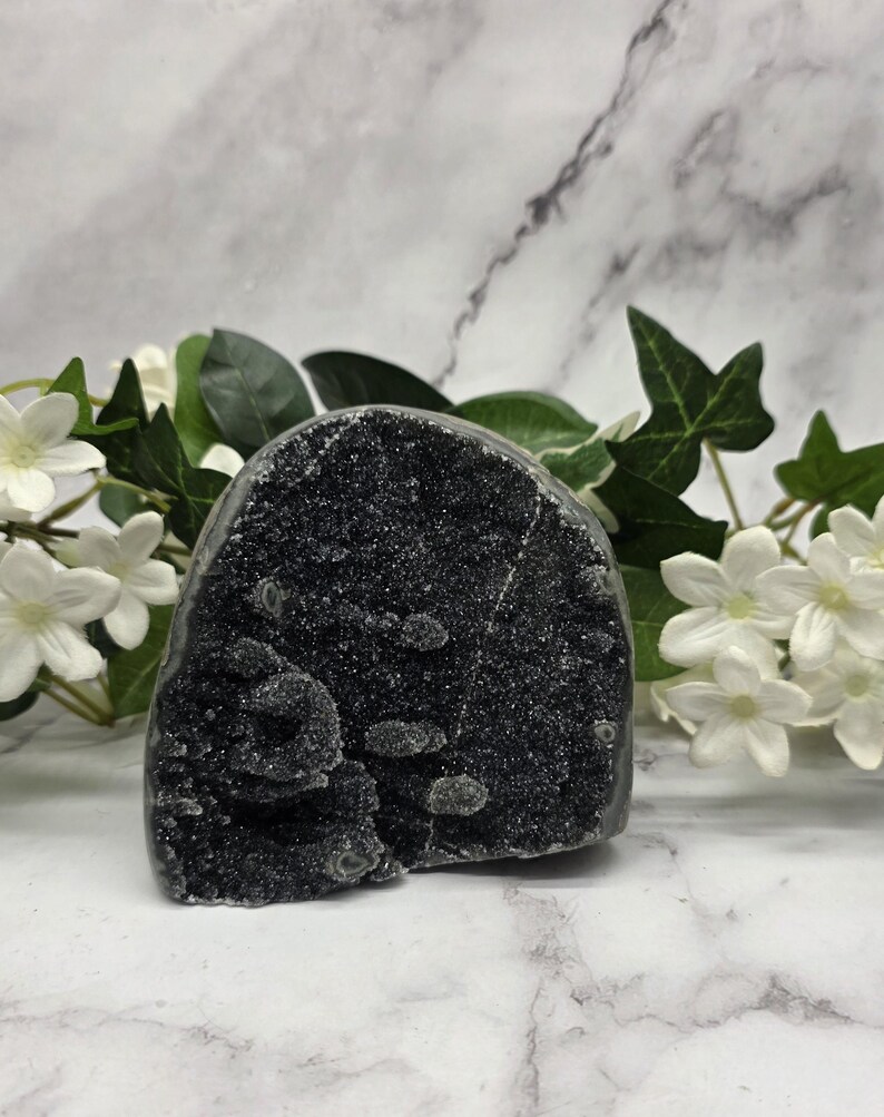May include: A dark gray crystal geode with a sparkling, textured surface. The geode has a rounded shape with a rough, uneven texture. White flowers and green leaves are arranged around the geode.