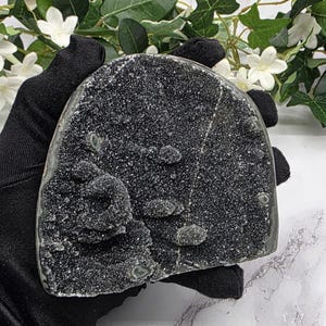 May include: A dark gray crystal geode with a sparkling, textured surface. The geode is held by a gloved hand, with white flowers and green leaves in the background. The crystal has a rounded shape.