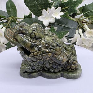 May include: A dark green stone sculpture of a three-legged toad with a textured, bumpy back and a round coin held in its mouth. The toad is depicted in a seated position on a flat, irregular base, featuring carved details on its skin and face.