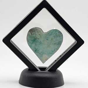 May include: A heart-shaped green and white stone displayed in a black diamond-shaped display stand.