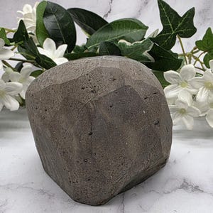 May include: A dark gray, faceted stone sculpture with a rough, textured surface. The sculpture is a geometric shape, possibly a decorative object or paperweight. White flowers and green leaves are in the background.