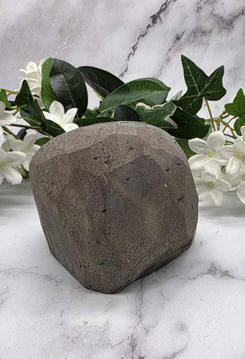May include: A dark gray, faceted stone sculpture with a rough, textured surface. The sculpture is a geometric shape, possibly a decorative object or paperweight. White flowers and green leaves are in the background.