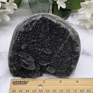 May include: A dark, geode-like stone with a rough, textured surface. The interior is filled with sparkling black crystals. A ruler is placed next to the stone for scale. The stone is approximately 4 inches wide.