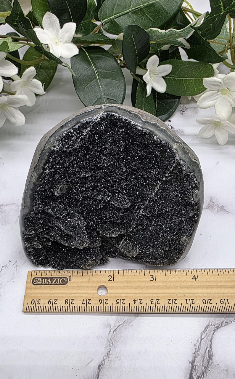 May include: A dark, geode-like stone with a rough, textured surface. The interior is filled with sparkling black crystals. A ruler is placed next to the stone for scale. The stone is approximately 4 inches wide.