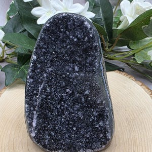May include: A dark gray geode crystal with a sparkling, textured surface. The crystal is displayed on a wooden slice, with white flowers and green leaves in the background. The geode has a rounded, irregular shape.