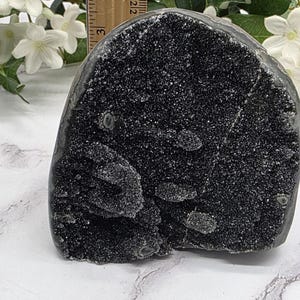 May include: A dark, sparkling black crystal geode, approximately 3 inches tall, with a rough, textured surface. The geode is displayed on a marble-patterned surface, with white flowers and greenery in the background.