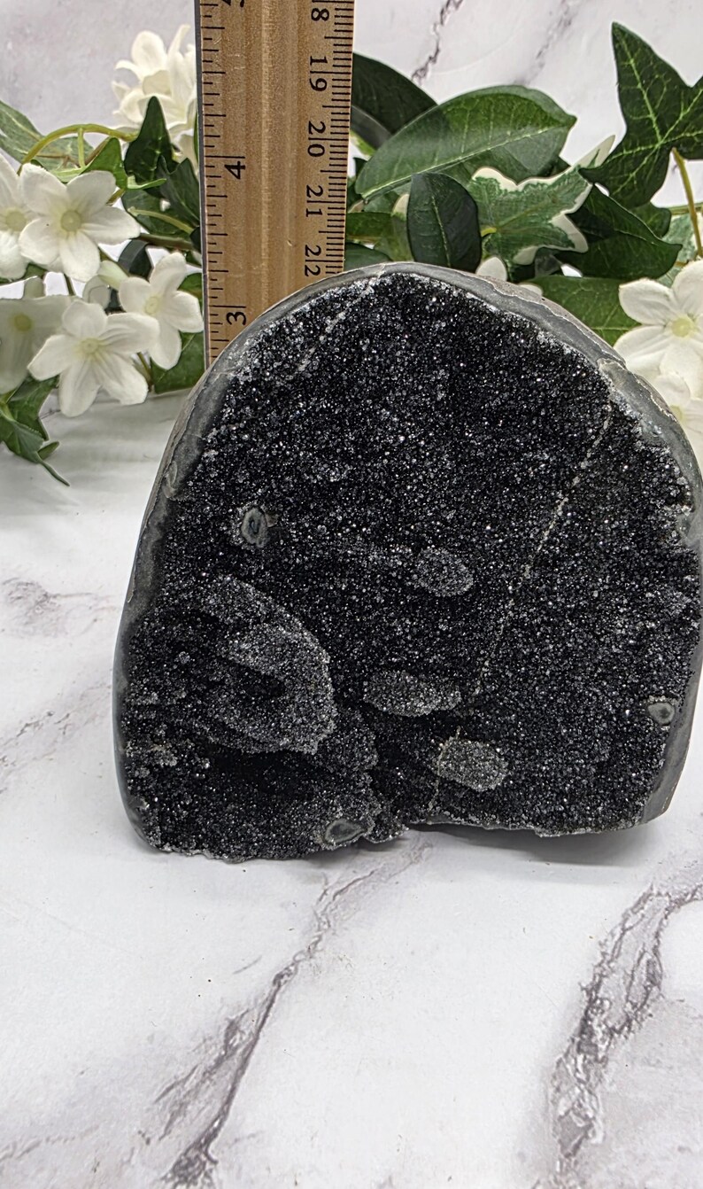 May include: A dark, sparkling black crystal geode, approximately 3 inches tall, with a rough, textured surface. The geode is displayed on a marble-patterned surface, with white flowers and greenery in the background.
