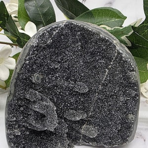 May include: A dark, textured geode with a sparkling, crystalline surface. The geode is a deep black color and has a rough, uneven shape. Green leaves and white flowers are in the background.