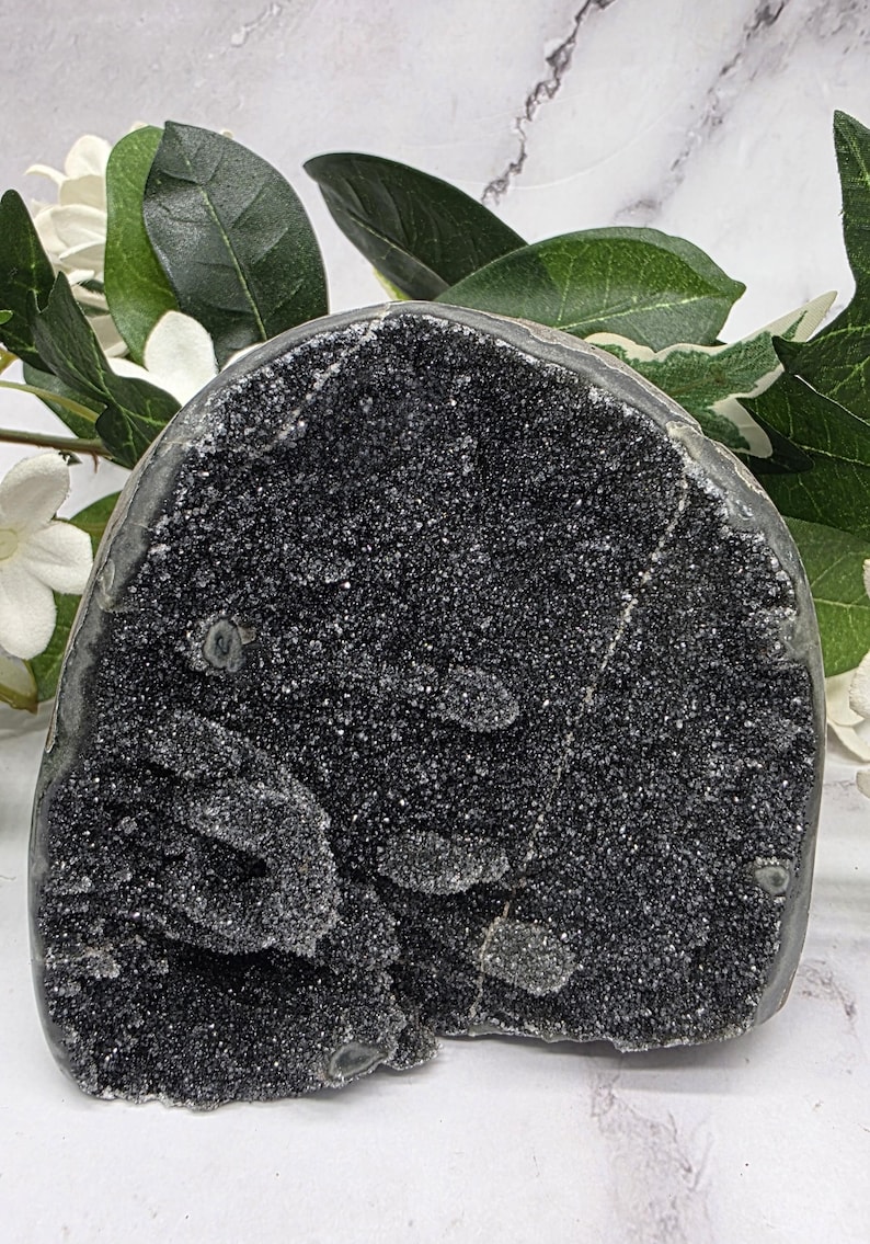 May include: A dark, textured geode with a sparkling, crystalline surface. The geode is a deep black color and has a rough, uneven shape. Green leaves and white flowers are in the background.