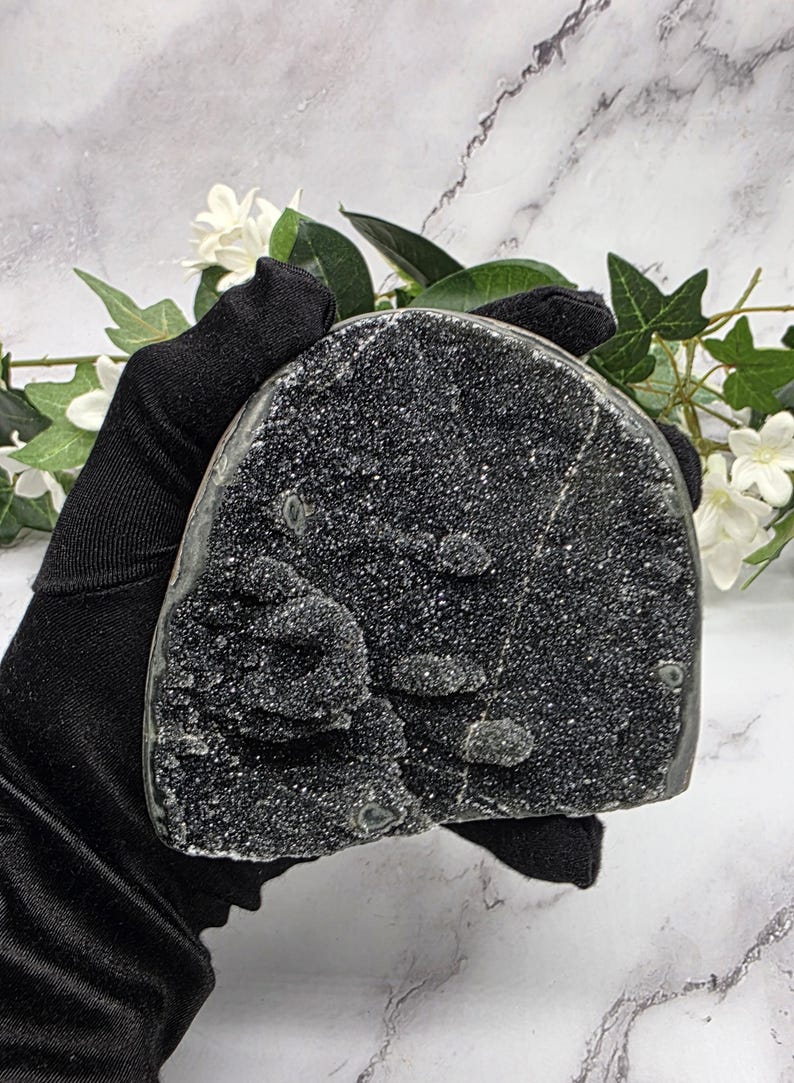 May include: A dark, sparkling black geode crystal with a rough, textured surface. The crystal is held in a black gloved hand, with white flowers and green leaves in the background. The geode has a rounded shape.