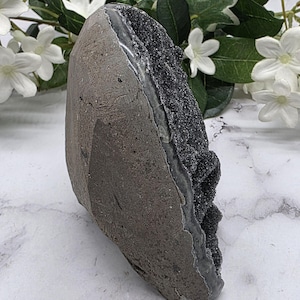 May include: A dark gray geode with a sparkling black crystal interior. The geode has a rough, textured exterior and a smooth, polished interior. The geode is displayed on a white marble surface with white flowers and green leaves in the background.
