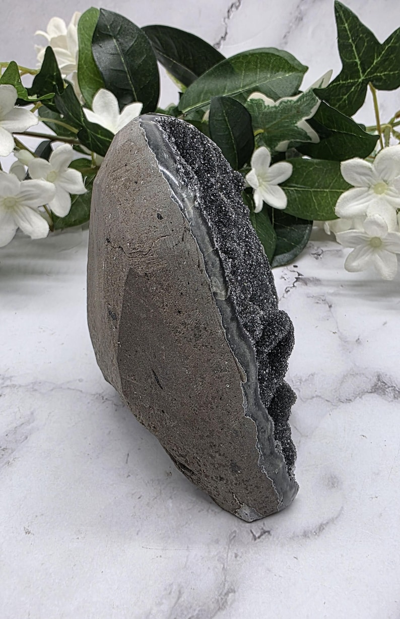 May include: A dark gray geode with a sparkling black crystal interior. The geode has a rough, textured exterior and a smooth, polished interior. The geode is displayed on a white marble surface with white flowers and green leaves in the background.