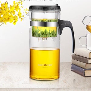 May include: A clear glass tea infuser with a black handle and lid. The infuser has a stainless steel band and contains green tea leaves. The bottom of the infuser is filled with amber-colored tea. A glass cup and books are in the background.