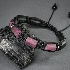 Peut inclure: Bracelet fait main avec des pierres précieuses roses rectangulaires et des perles noires tissées dans un cordon noir. Le bracelet est réglable avec une fermeture coulissante et des perles noires. Affiché sur une surface grise foncée.